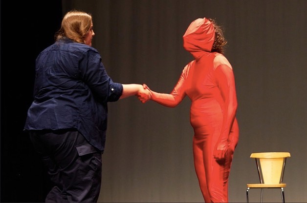 Two performers, one in a red morph suit shaking hands.