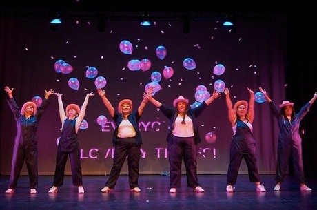 Six performers wearing hats and with their arms raised surrounded by balloons.
