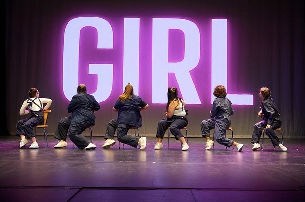 6 performers sat on chairs in front of large pink 'Girl' facing away from audience.