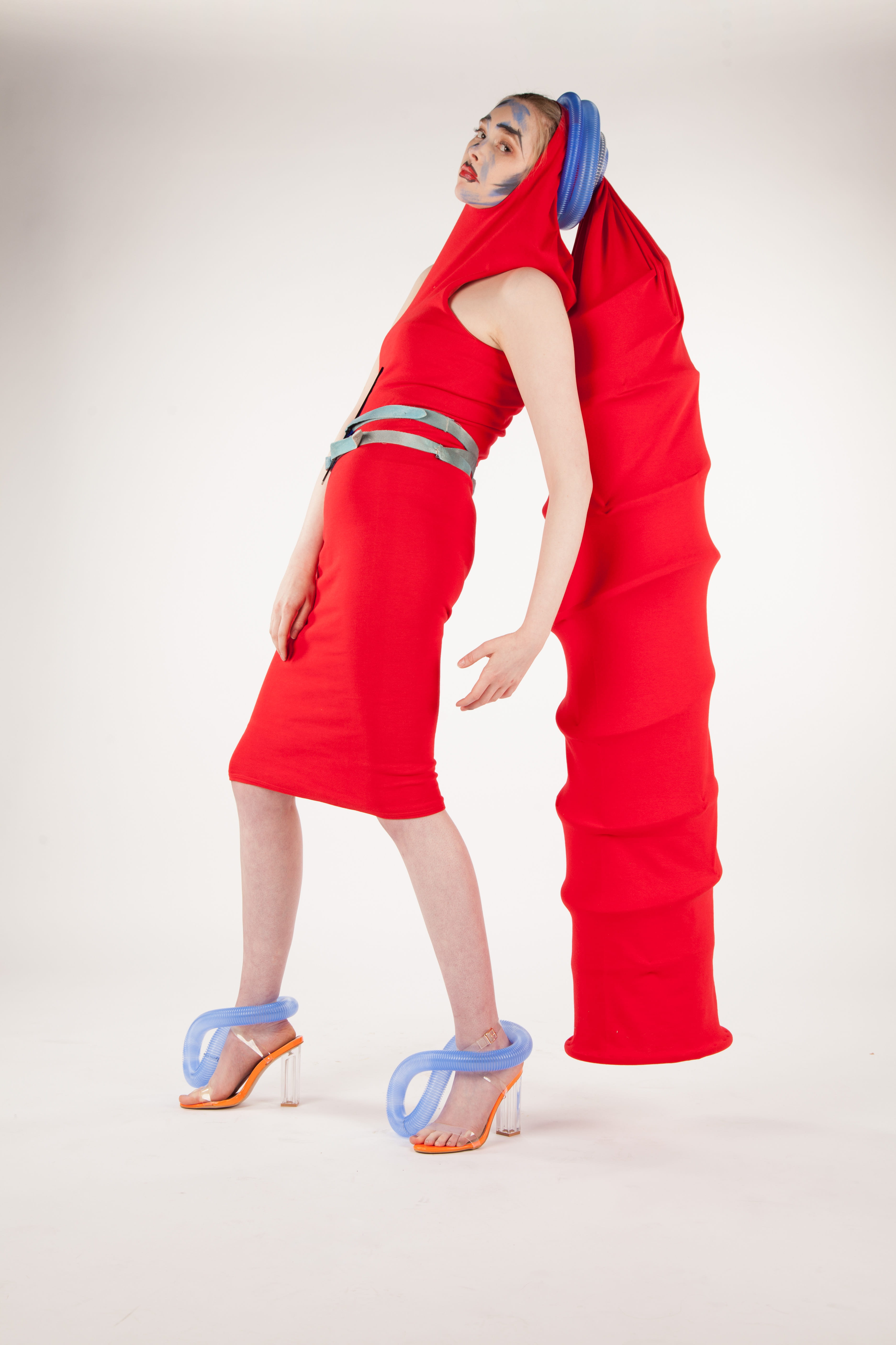 A female model in a red dress, with long, tubular head-wear hanging behind her.