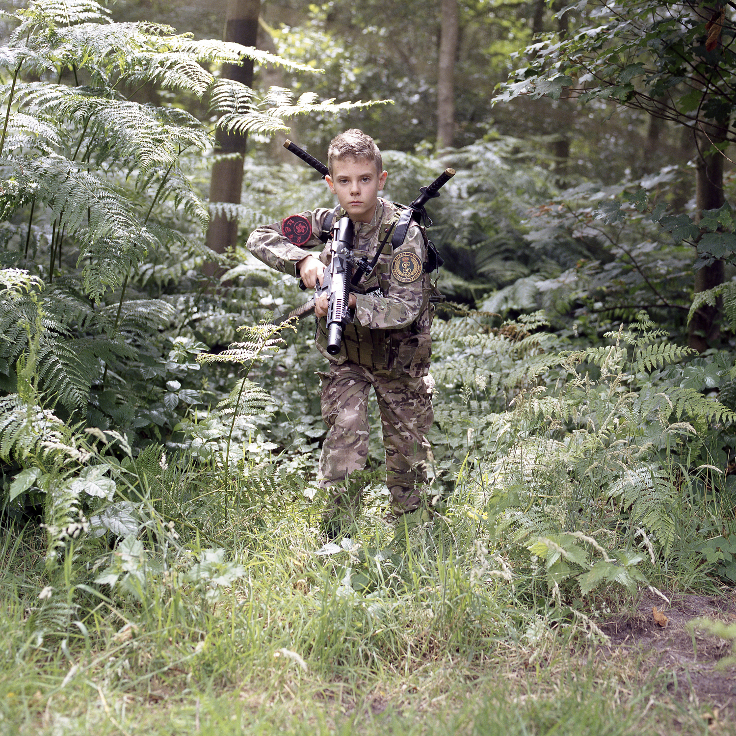 A young boy with shaved hair stands amongst several large bushes, appearing as if in a jungle. They are holding a small toy smg in one hand and are staring defiantly into the camera.