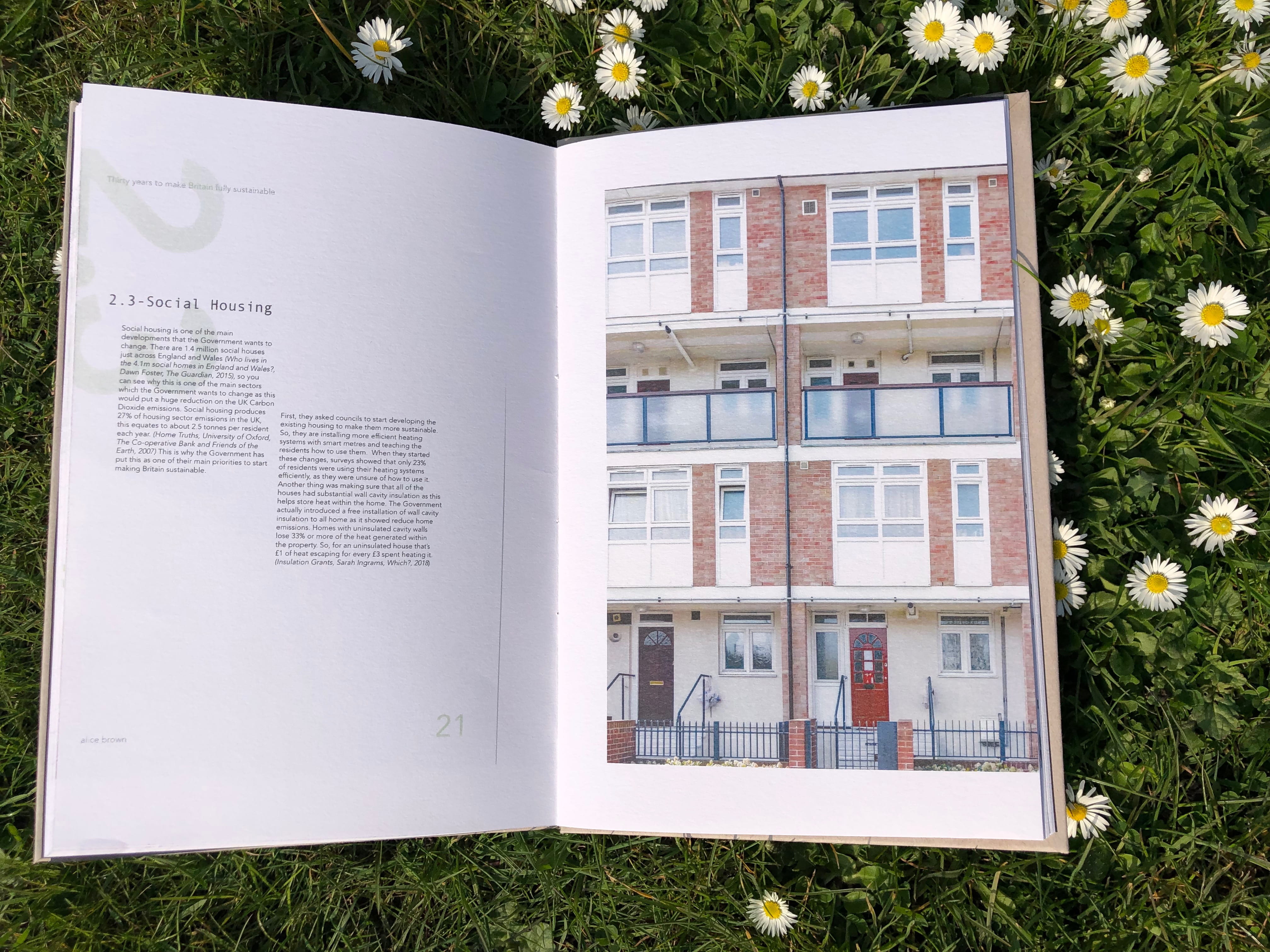 A book is laid on a patch of daisies, an image of a block of flats is shown on one page, the accompanying page is titled 