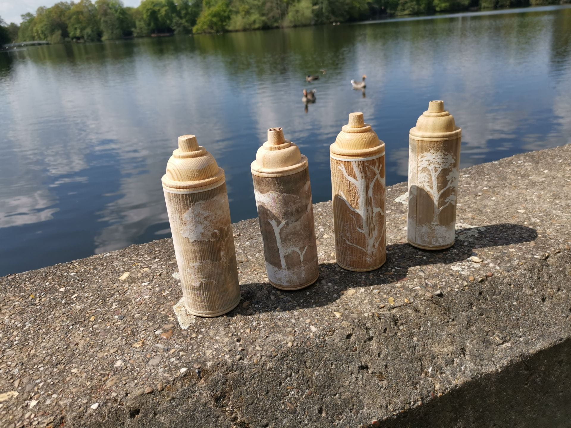 Four wooden spray paint cans by a lakeside, images of trees engraved into them.