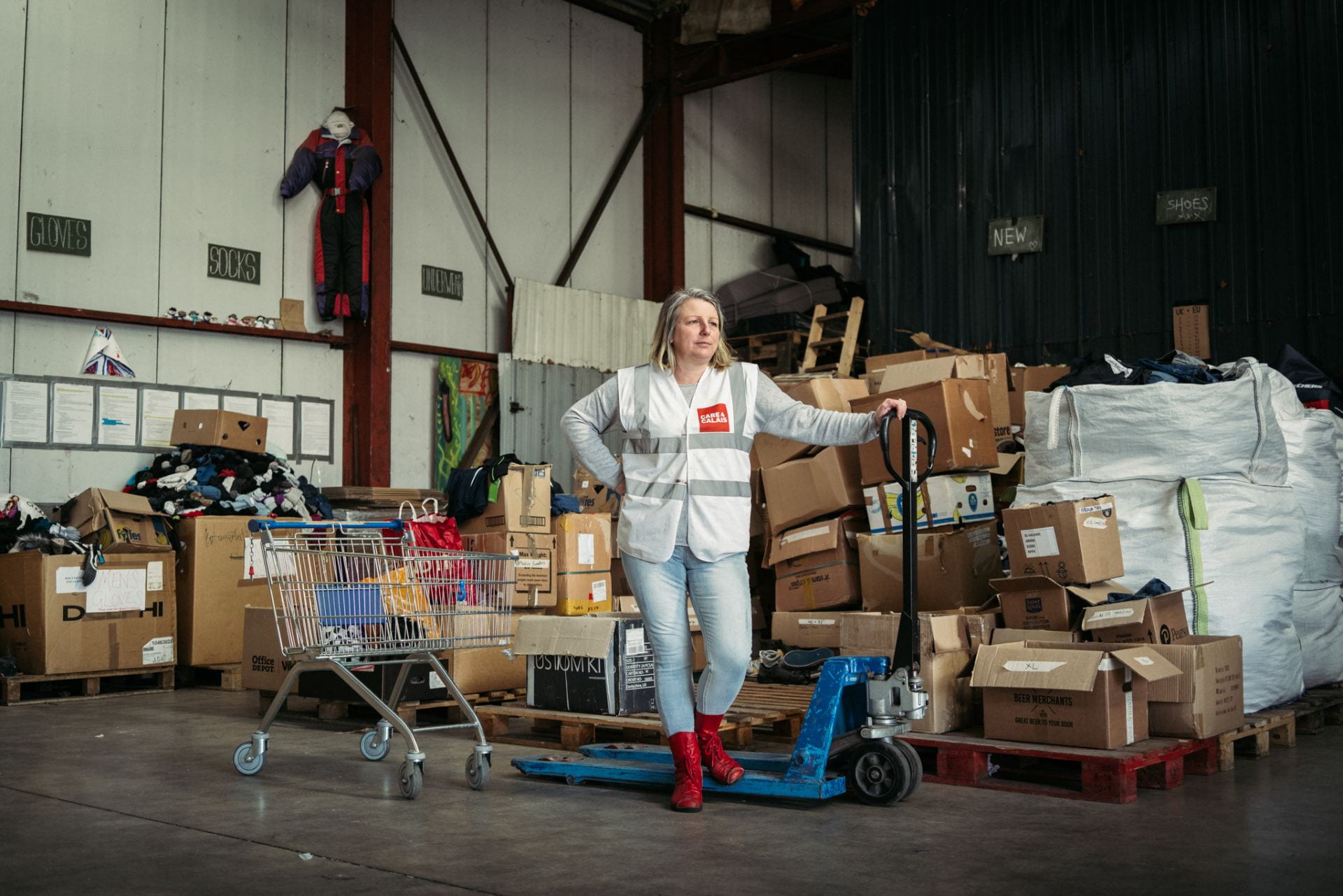 Volunteer with red shoes and off-white high visibility jacket standing next to a trolley, inside warehouse full of cardboard boxes containing clothes.