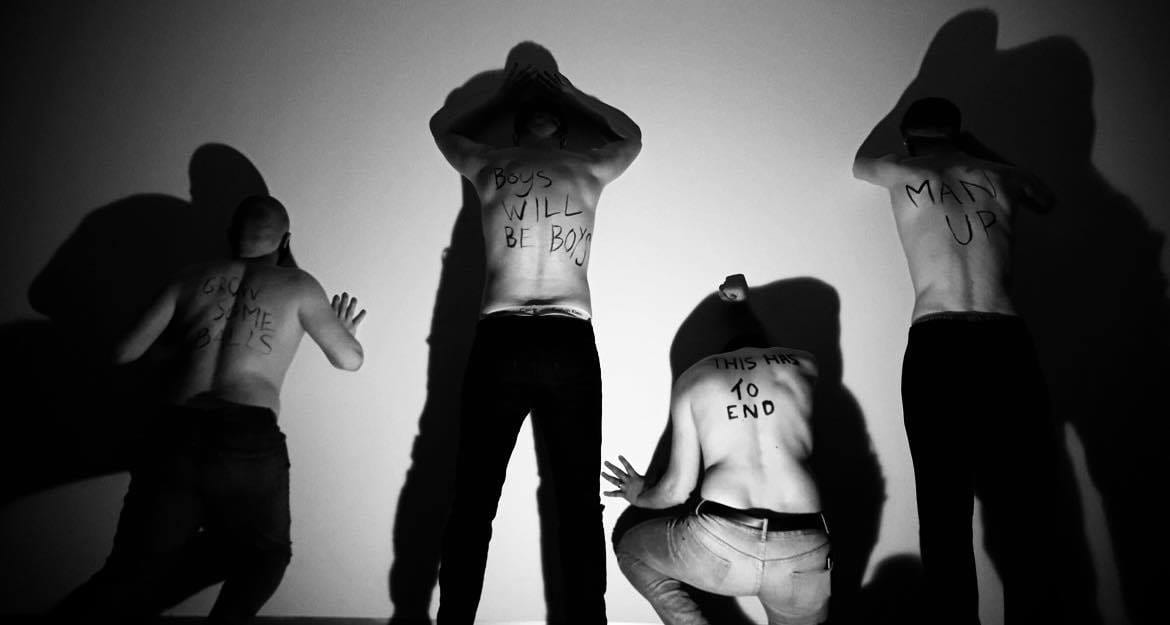 4 men stood pushing against a wall in a line, they have writing on their backs which include 