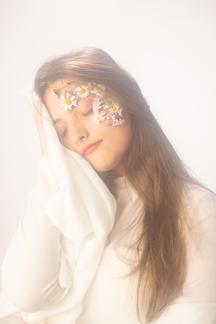 Image of a women wrapped in a white cloth with flowers on her face. The lighting is ethereal and light. It is a close up portrait.
