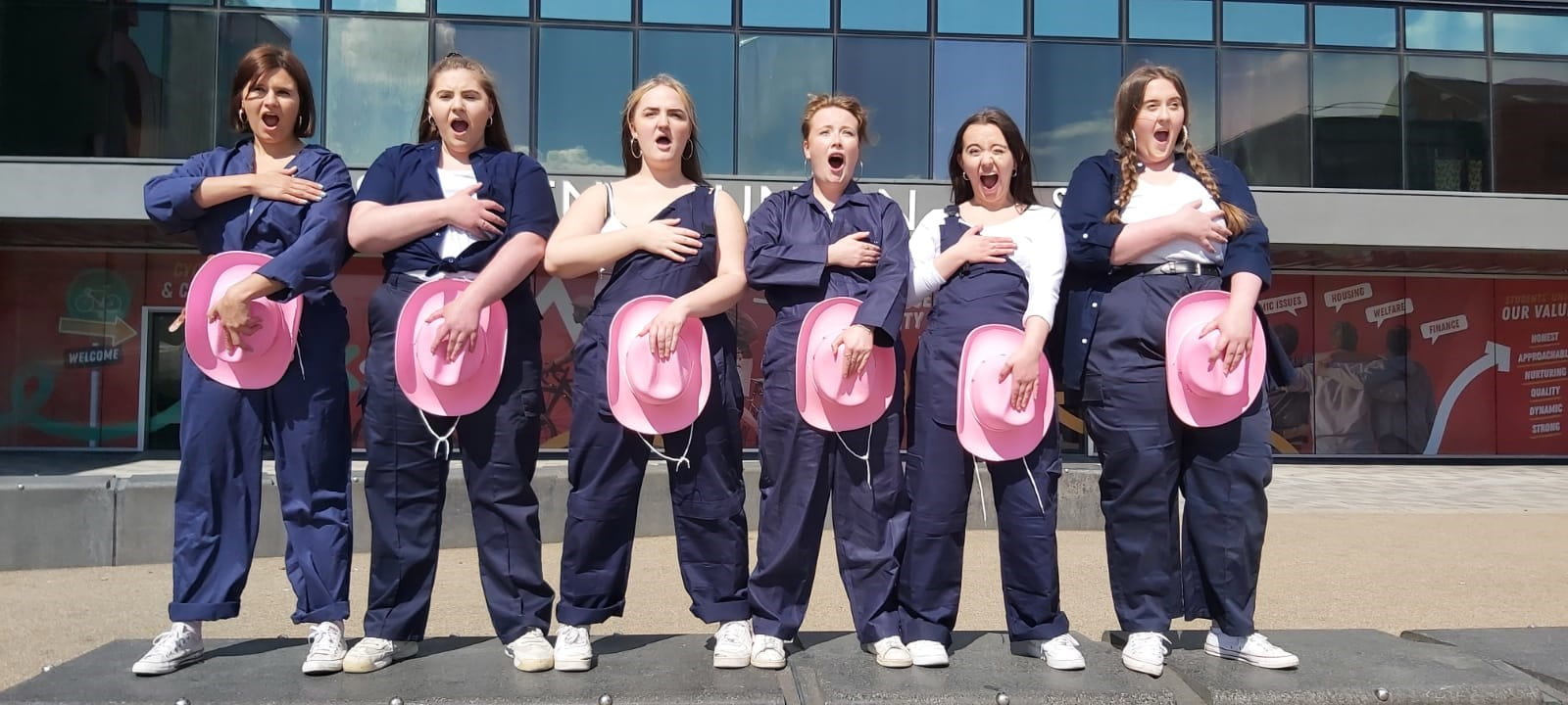 Image contains six women holding pink cowboy hats over their crotch. All dressed in men's workwear.