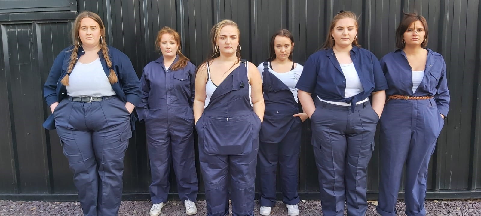 Image contains six women standing face on to the camera, looking serious. All dressed in men's workwear.