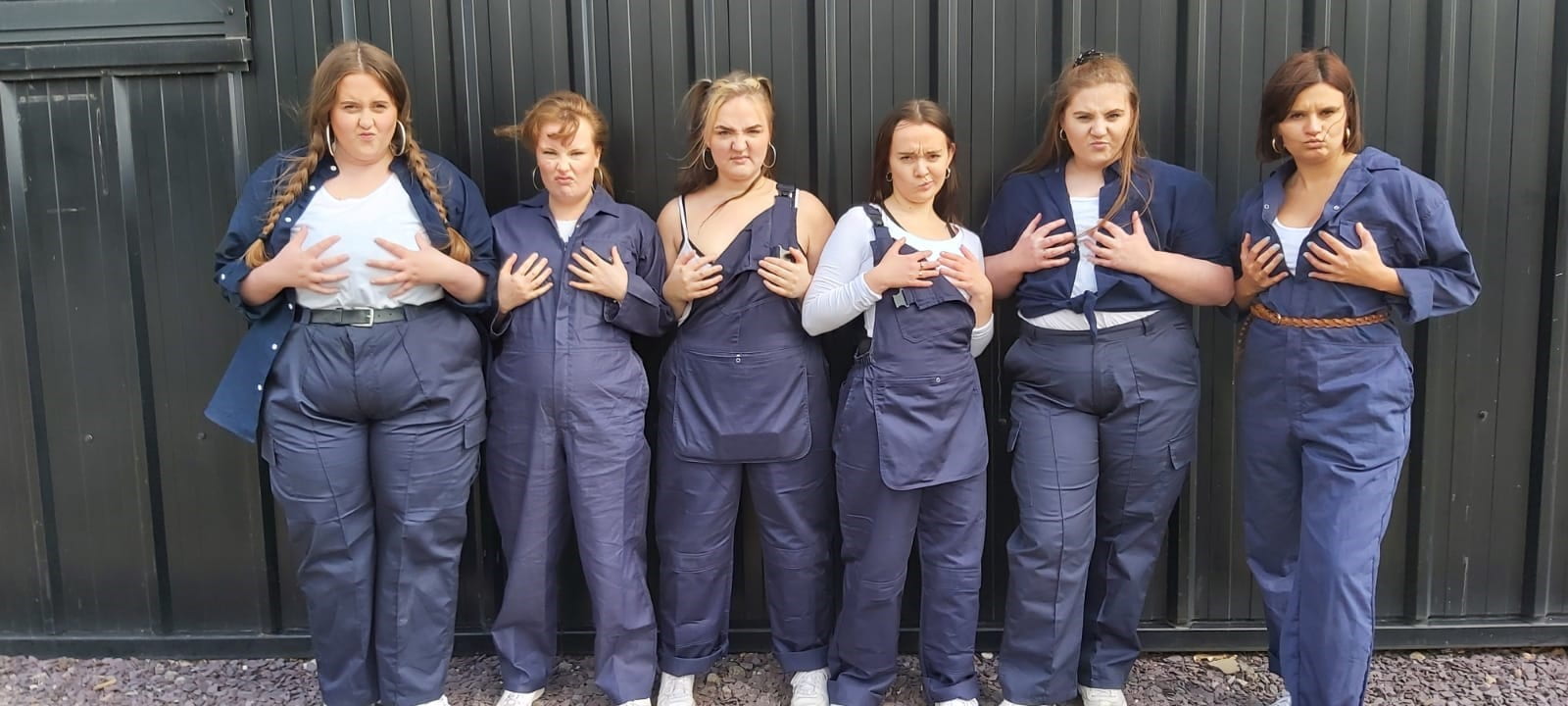 Image contains six women all holding their chest, with a scowl on their faces. All dressed in men's workwear.